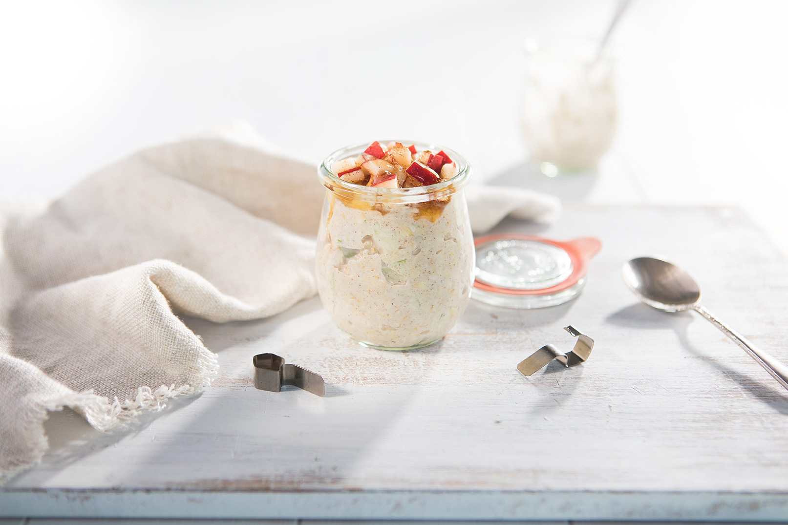 Image of apple pie overnight oats in a glass jar with the lid off on a white cutting board with a spoon and white cloth napkin on the side