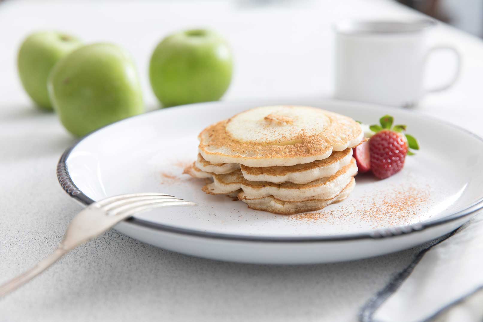 Stacked apple ring pancakes sprinkled with cinnamon on a large white plate with a strawberry and fork on the side for serving.