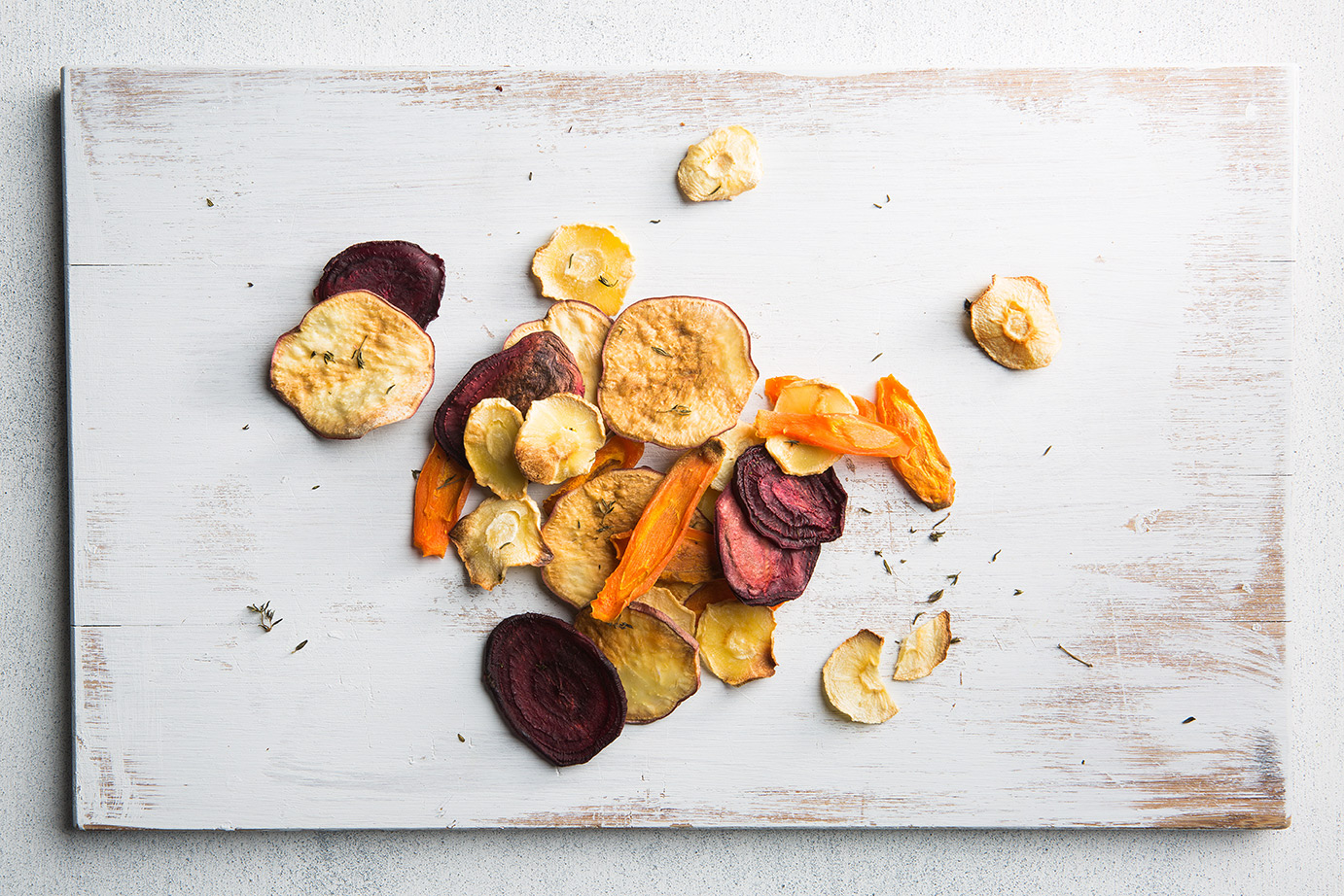 Image of baked root vegetable chips spread on a white wooden chopping board