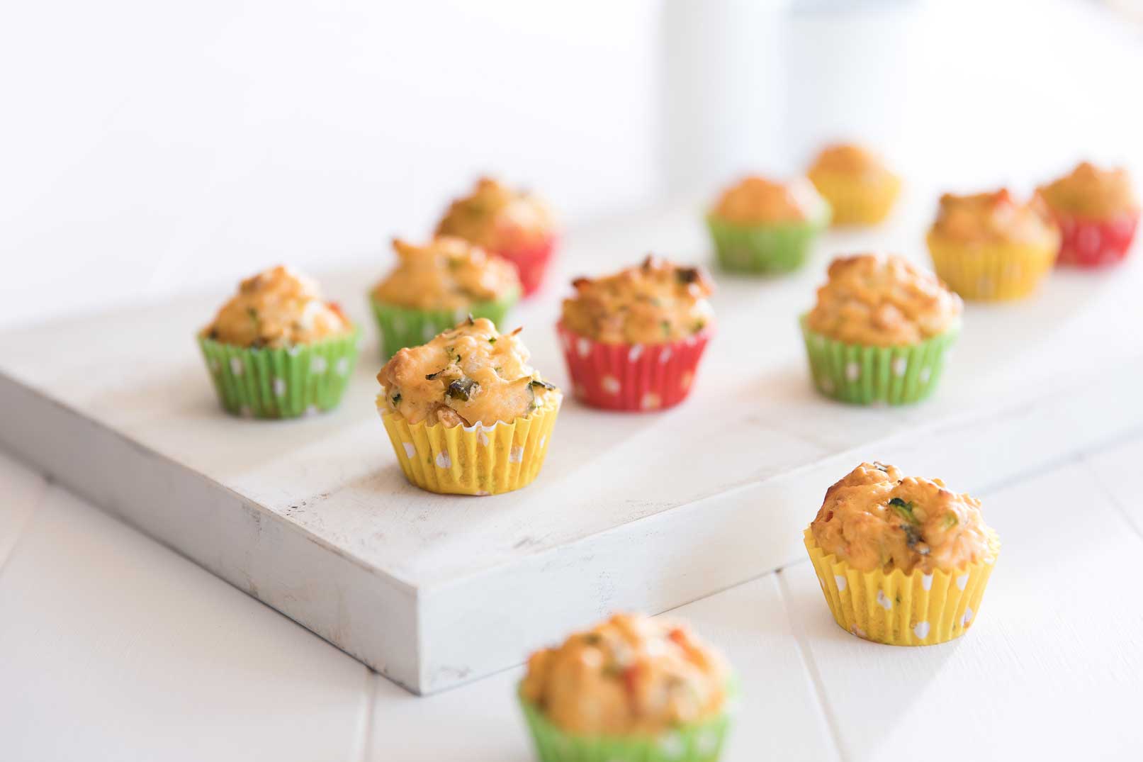 Baked bean mini muffins in yellow, red and green patty cases on a white cutting board.