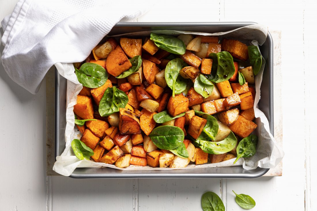 Roasted vegetables in a metal baking dish lined with baking paper, garnished with fresh spinach leaves
