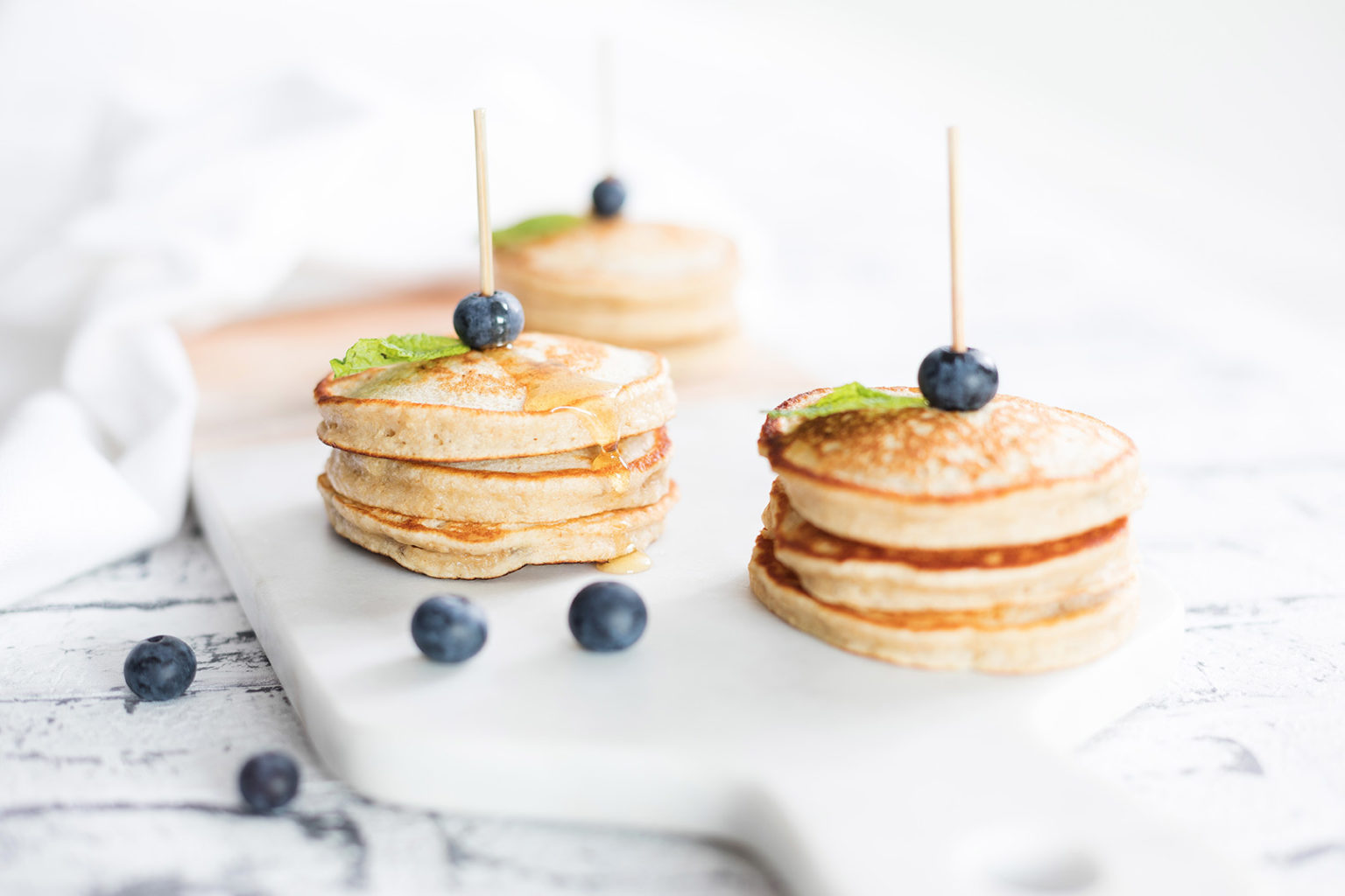 Three stacked piles of banana pikelets with a skewer and blueberries served on a chopping board