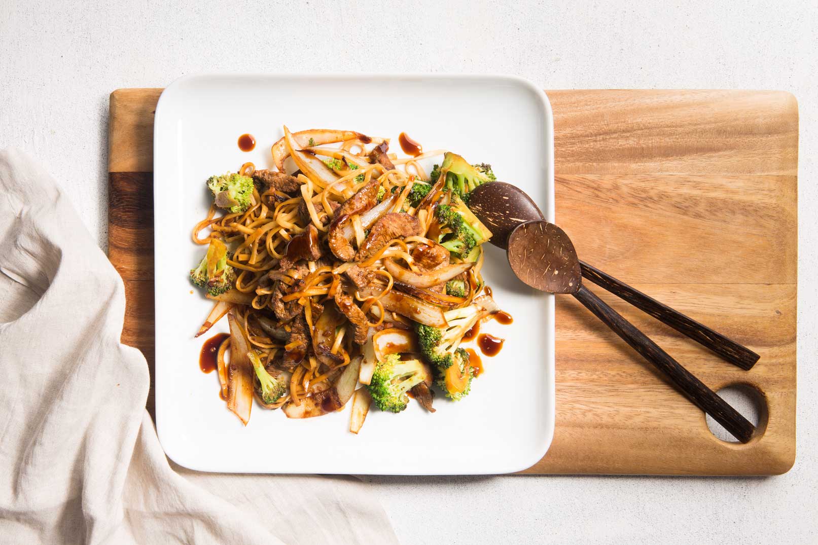 Image of beef and broccoli stir fry on a white square plate sitting on a wooden chopping board with wooden serving spoons.