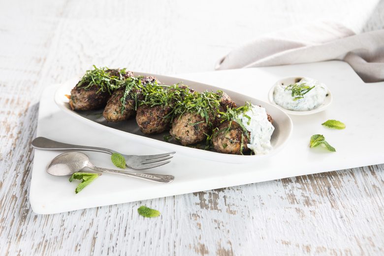 Image of five baked beef koftas on a long serving dish topped with Tzatziki and herbs with serving utensils on the side 