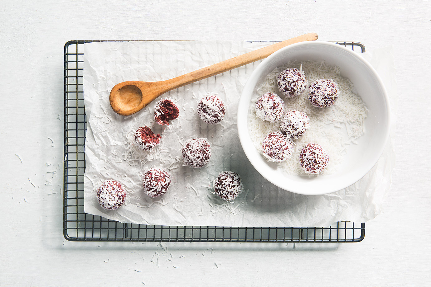 Image of BeetrootSpinacImage of six beetroot and spinach bliss balls in a bowl of coconut for rolling and another six balls on a baking paper on a cooling rack with a wooden spoon