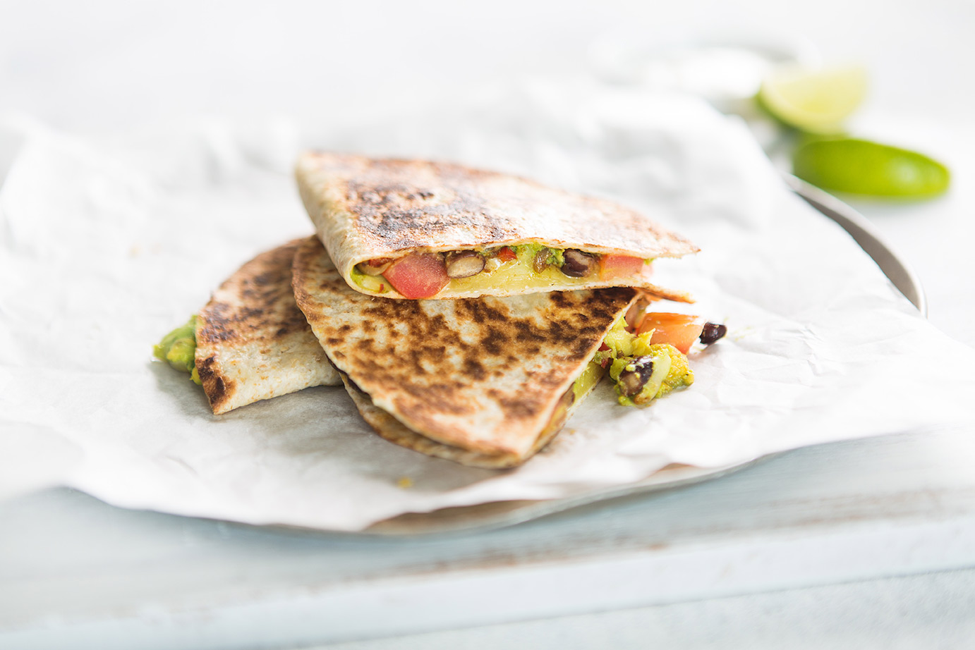 Image of three stacked blackbean quesadillas on baking paper on a round metal baking tray with sliced limes for serving in the background 