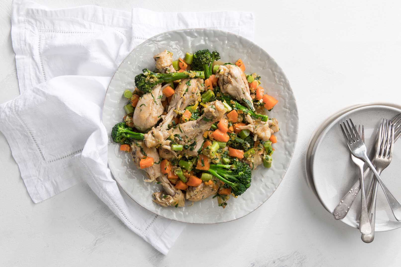 Cooked braised chicken and vegies in a large white plate with a white cloth napkin, serving plates and forks on the side.