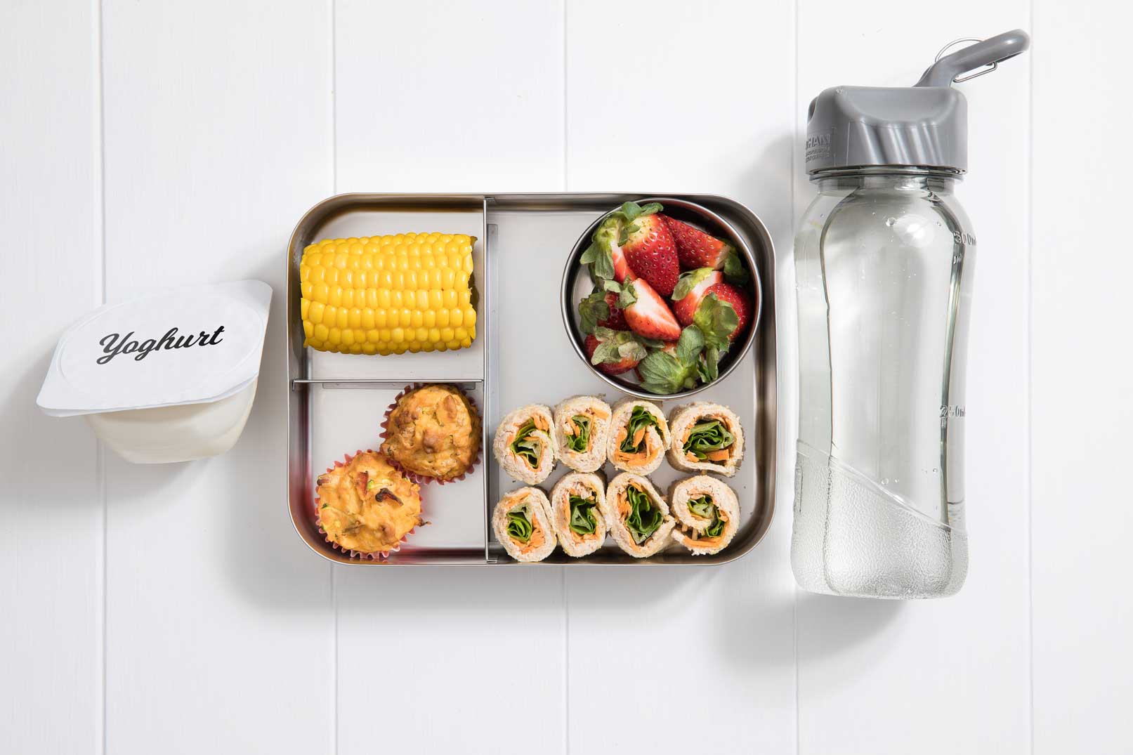 a compartment metal lunch box packed with eight sushi bread rolls, corn cob, small serving dish of strawberries, two baked bean muffins and a tub of yoghurt to the left and filled water bottle to the right.