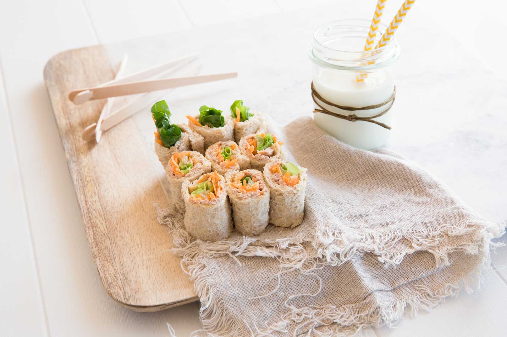 Nine bread sushi rolls served on a wooden chopping board with chopsticks and a jar of milk with two straws on the side.
