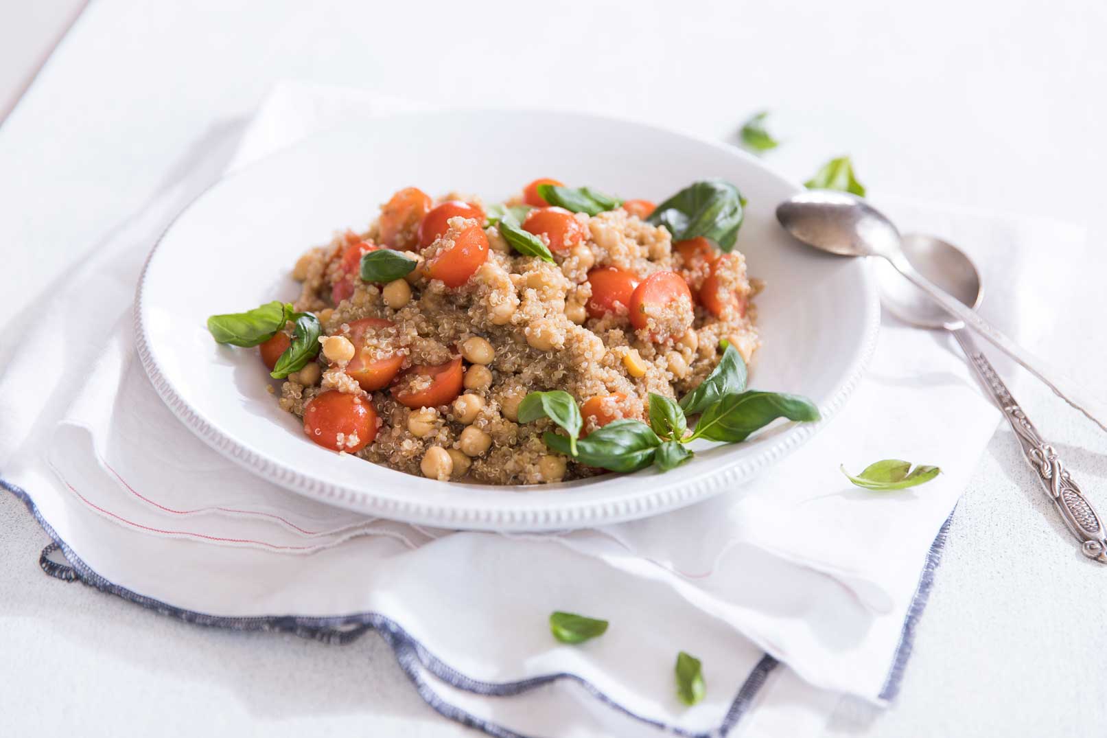 Caprese quinoa salad in a white bowl sitting on a white cloth napkin with blue trim and metal serving spoons on the side and a scattering of basil leaves.