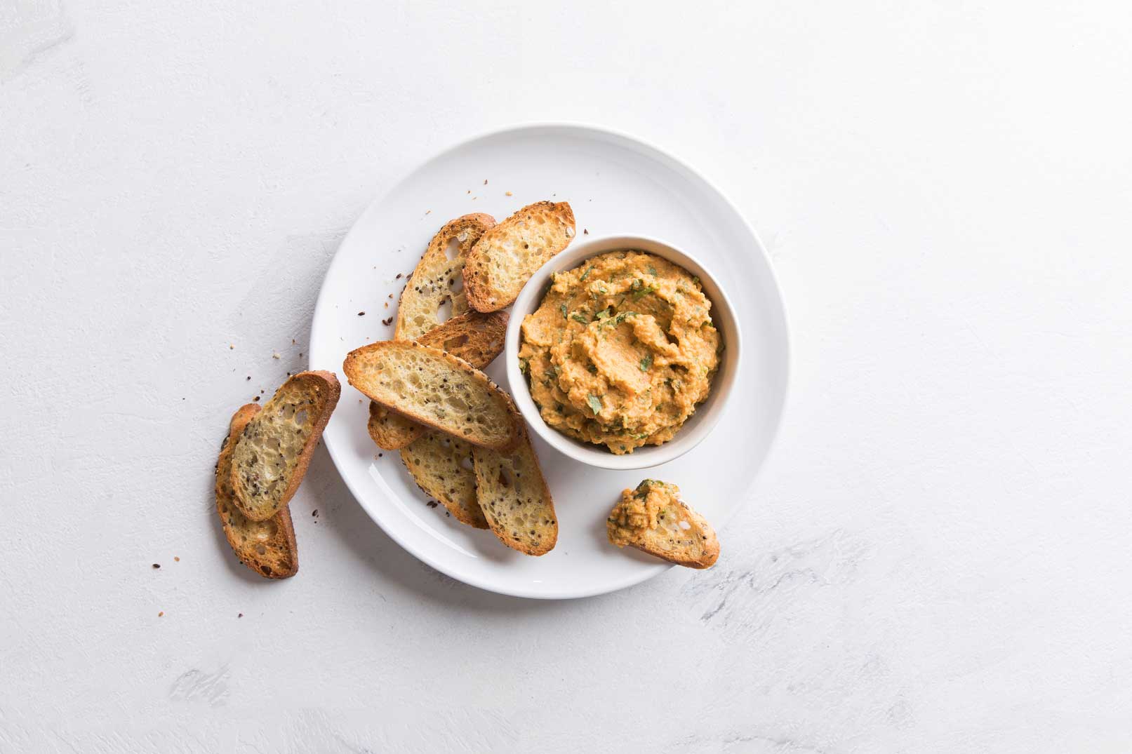 Carrot and ricotta dip in a small white bowl served on a white plate with baked bread slices for serving.