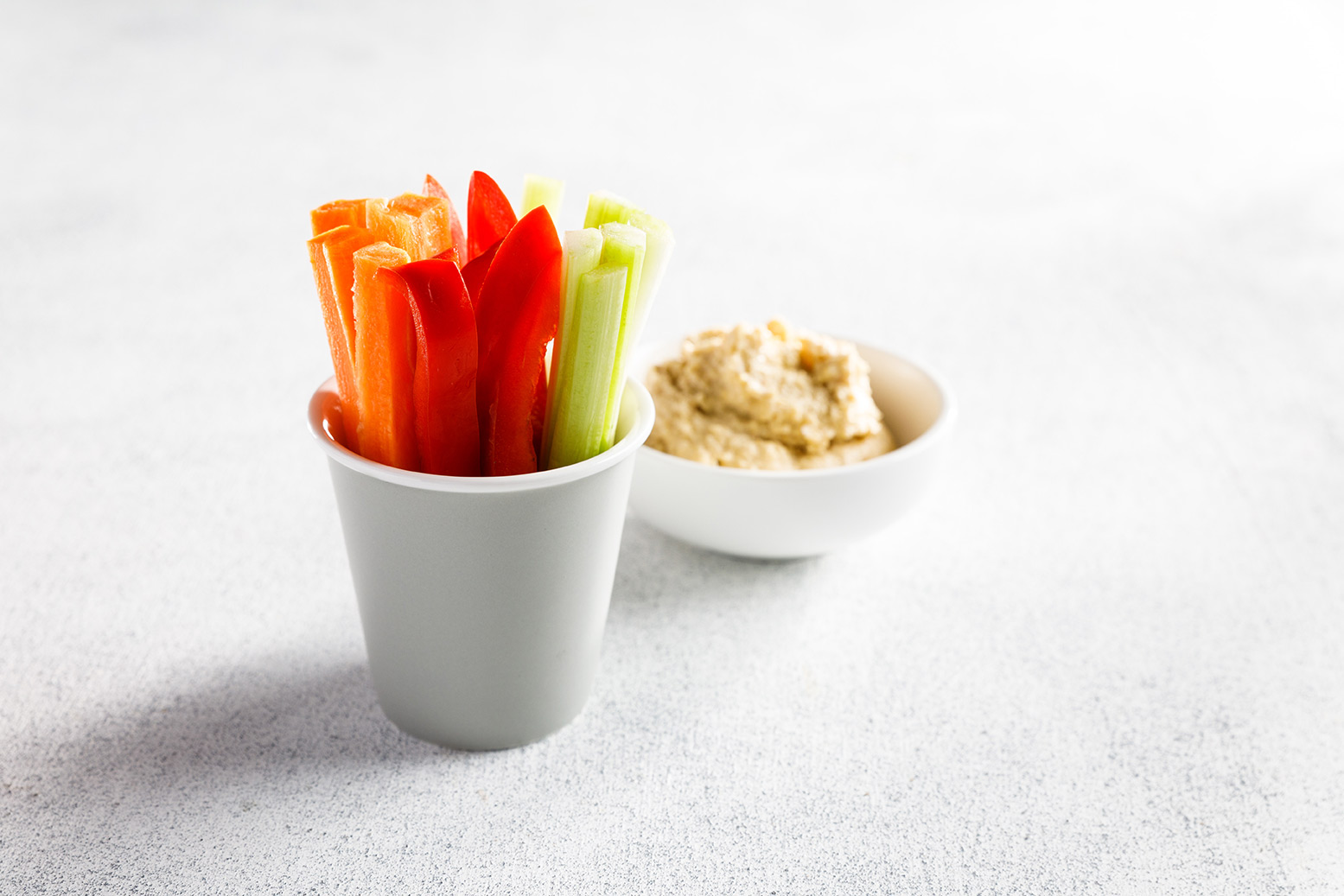 Image of carrot celery and red capsicum sticks standing in a white serving cup with hummus in a small white bowl on the side