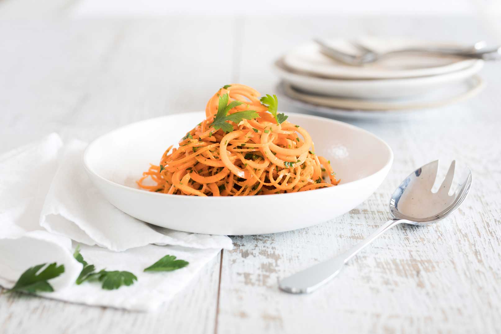 Carrot salad with orange and ginger piled in a white serving bowl with a large serving spoon and a white cloth napkin on the side with serving plates and utensils in the background.