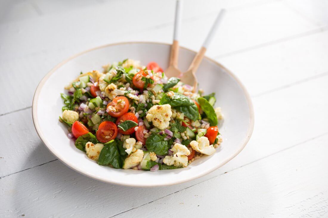 Cauliflower and barley salad in a large white bowl with a fork and spoon for serving on the side.