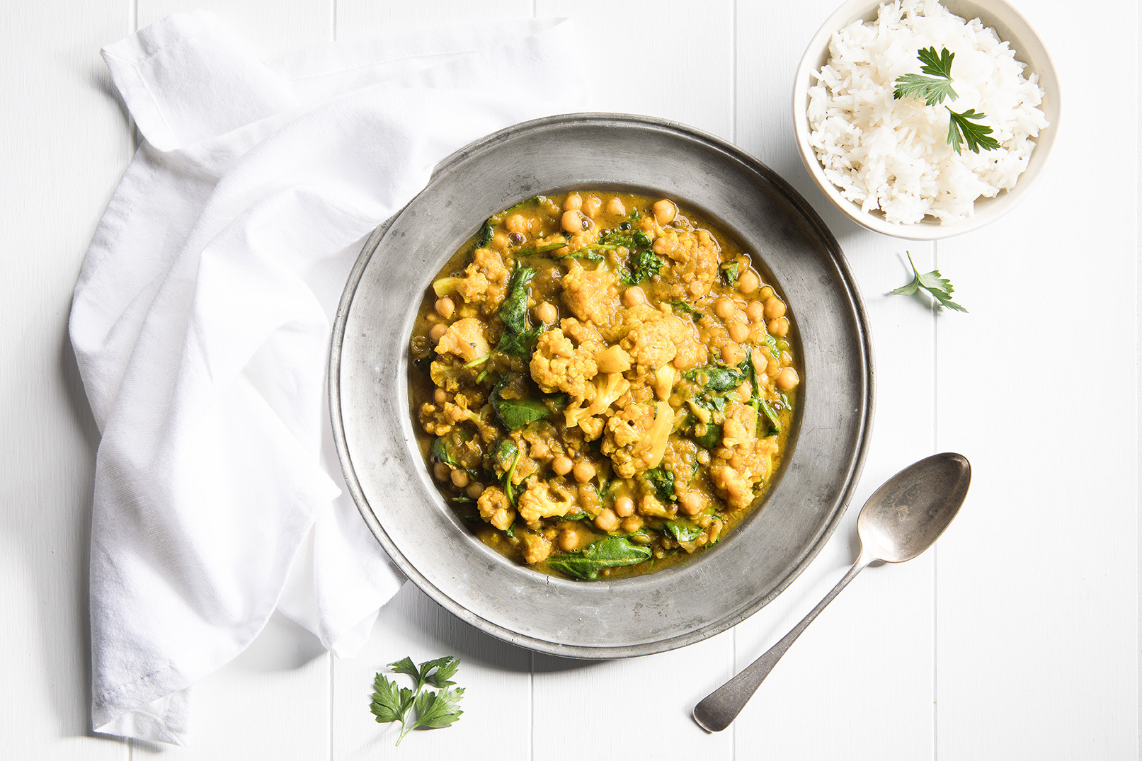 Image of cauliflower and spinach dahl in a large silver bowl with a metal spoon for serving and rice in a small white bowl on the side.
