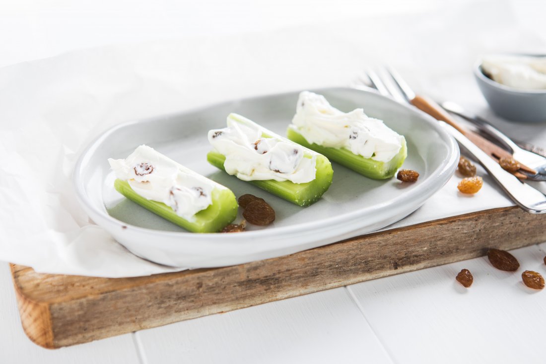 3 celery sticks spread with cream cheese and topped with sultanas on a white plate