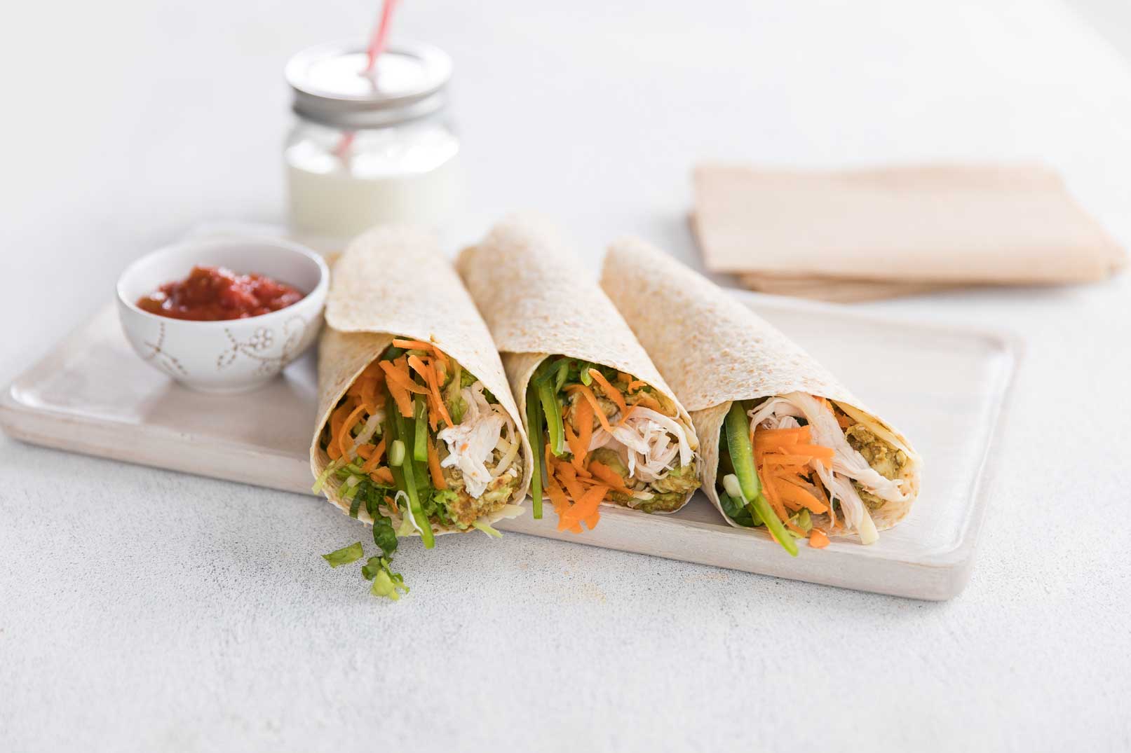 Three chicken fajita roll-ups served on a white cutting board with a small bowl of sauced on the side. A glass jar of milk with a straw and brown paper napkins are in the background.