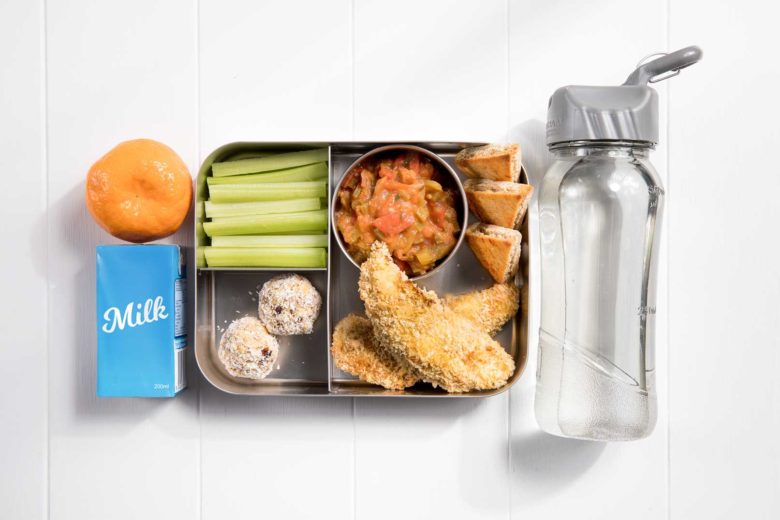 a compartment metal lunch box packed with two chicken fingers with a small serving dish of Mexican salsa and three pita bread triangles, celery sticks, two bliss balls and a mandarin and plain milk popper to the left and filled water bottle to the right