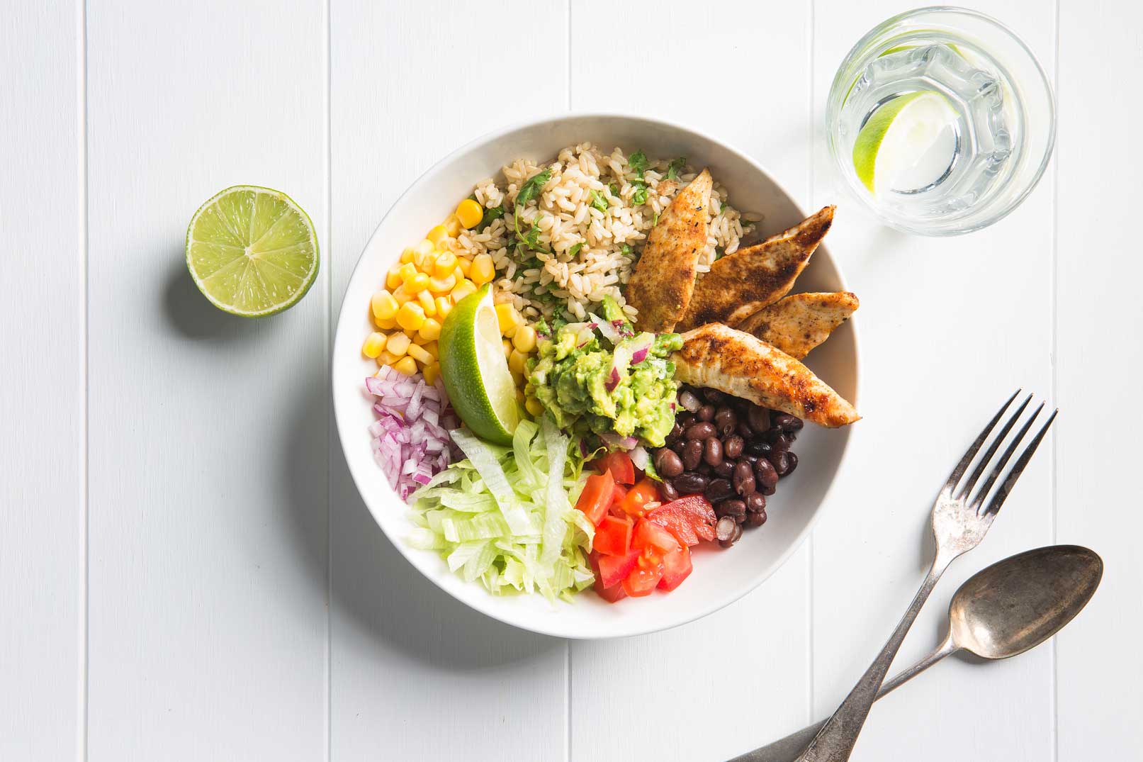 Image shot from above of a chicken burrito bowl with half a lime on the side, serving forks and a jug of water
