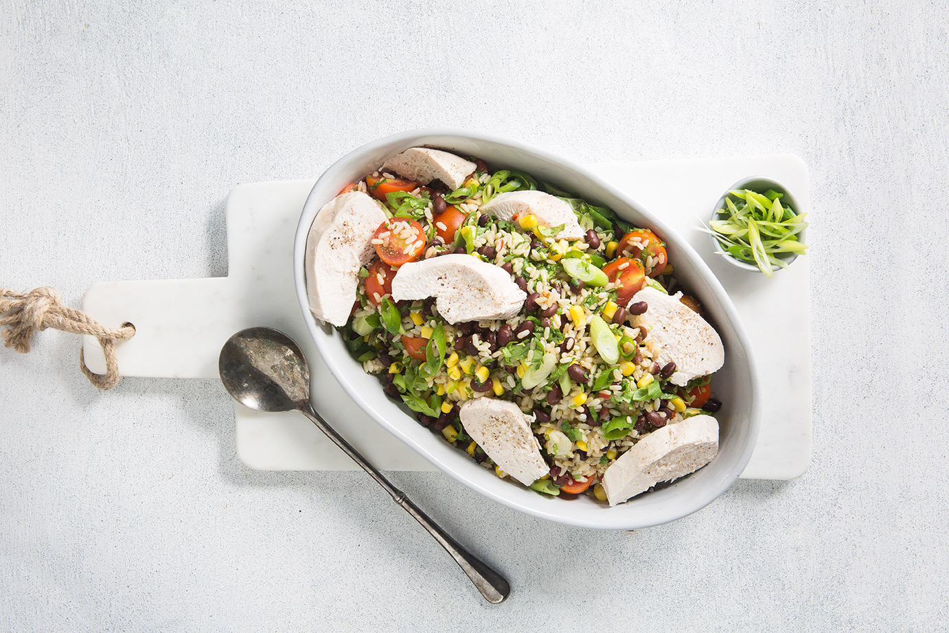 Image of chicken rice salad in a large white serving dish on a white cutting board shot from above with a large silver spoon for serving on the side