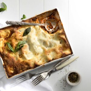 A baked chicken and vegetable lasagne with a metal serving spoon and a spoonful missing in a large rectangle oven proof dish, served with basil leaves, forks, dressing and a white cloth napkin on the side.