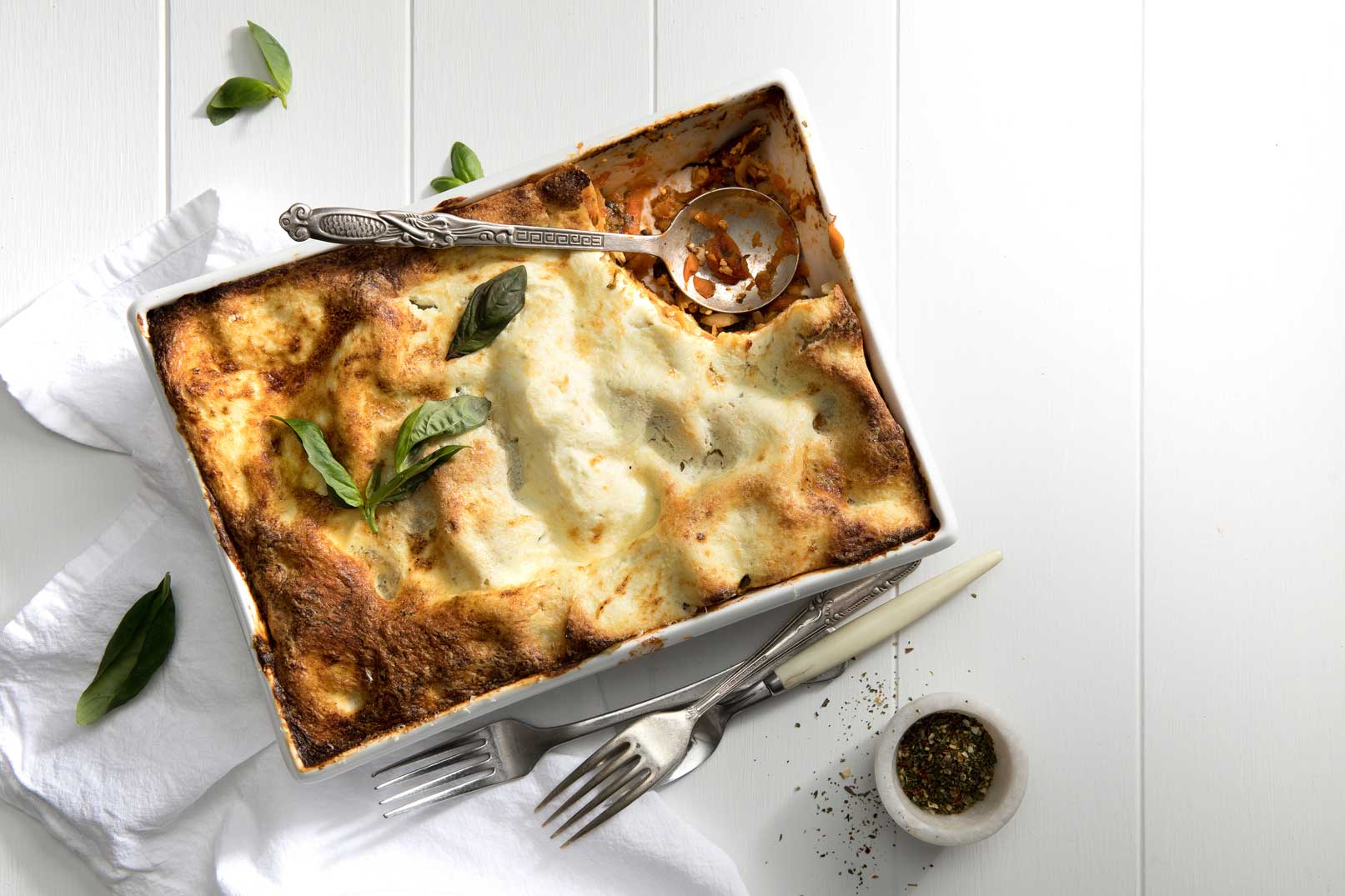 A baked chicken and vegetable lasagne with a metal serving spoon and a spoonful missing in a large rectangle oven proof dish, served with basil leaves, forks, dressing and a white cloth napkin on the side.