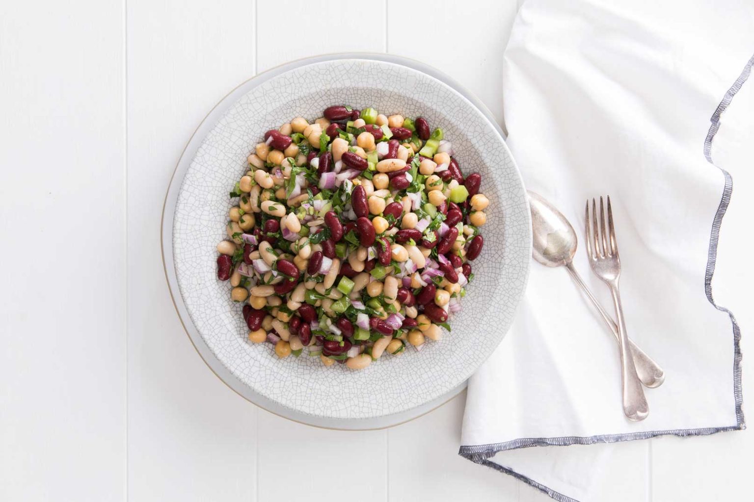 Classic bean salad in a large serving bowl with a metal spoon and fork and cloth napkin with blue trim on the side.