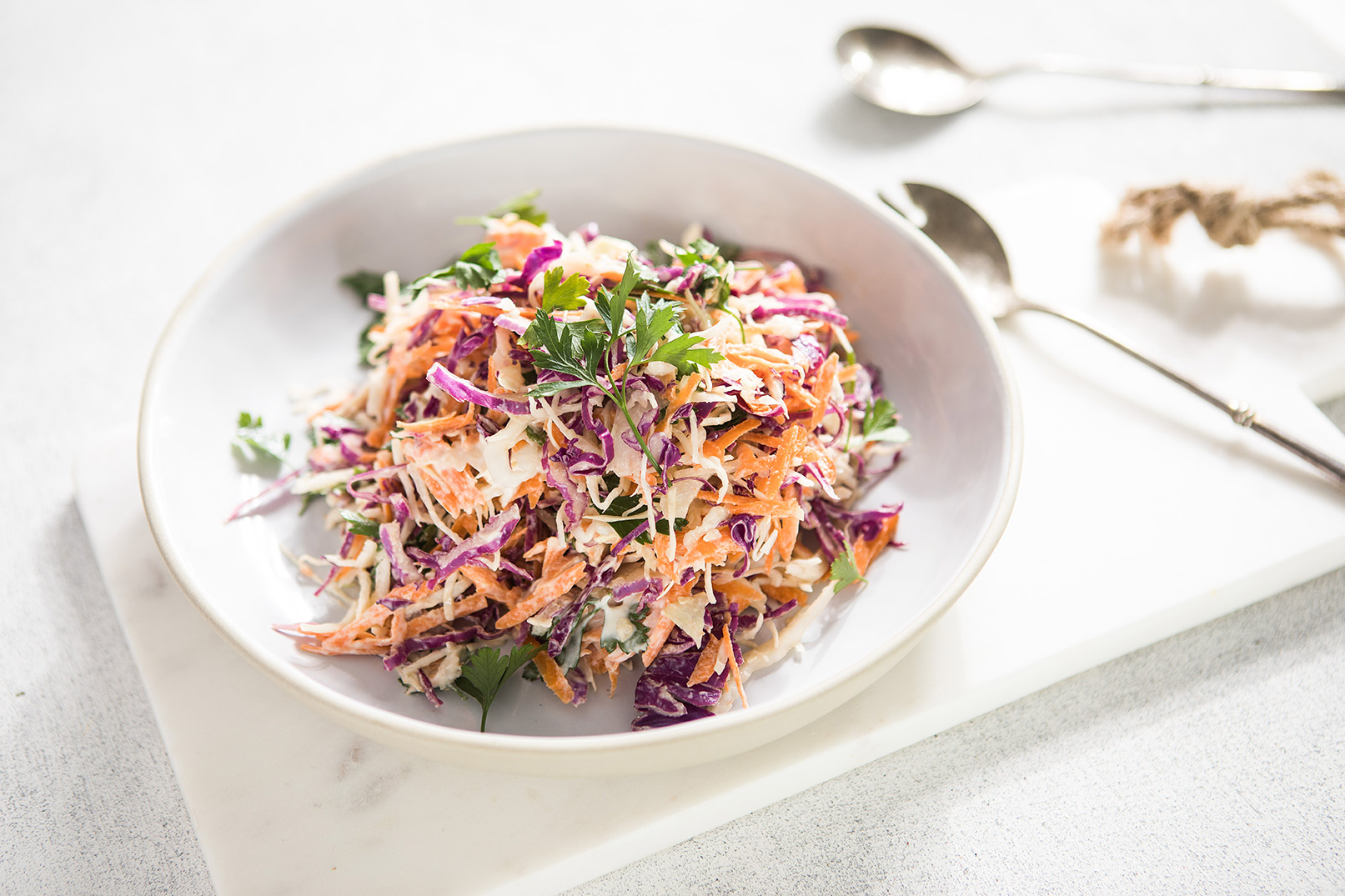 Image of coleslaw in a white bowl served on a white marble cutting board with serving utensils in the background