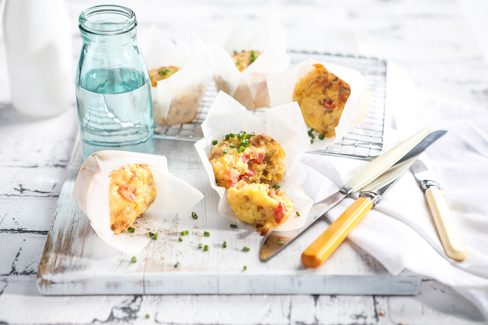 Image of baked easy savoury muffins in baking paper on a white cutting board and cooling rack with knives and a glass jug of water on the side. 