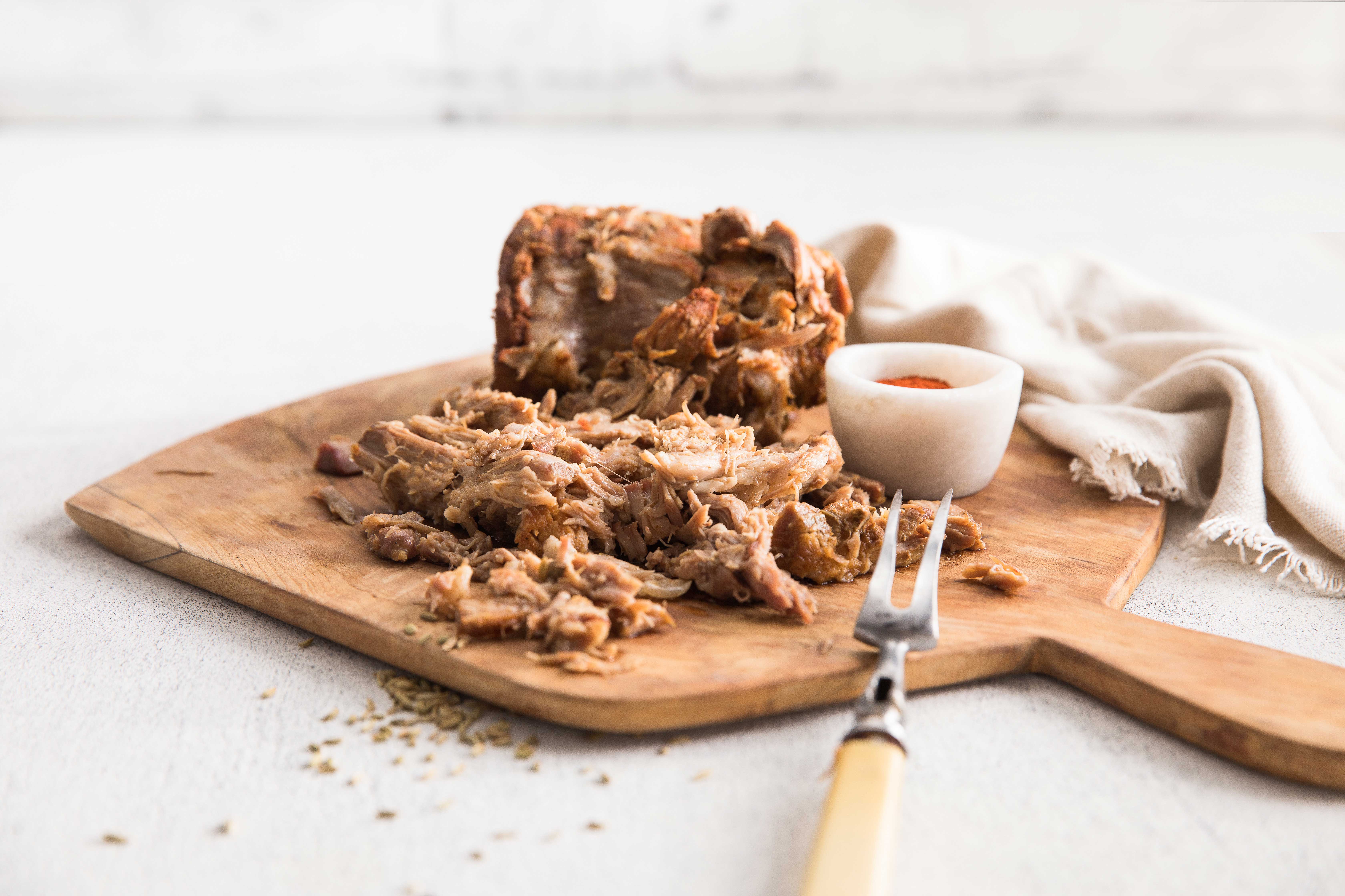 Easy peasy pulled pork shredded on a wooden cutting board with a large meat fork and a small bowl of sauce.