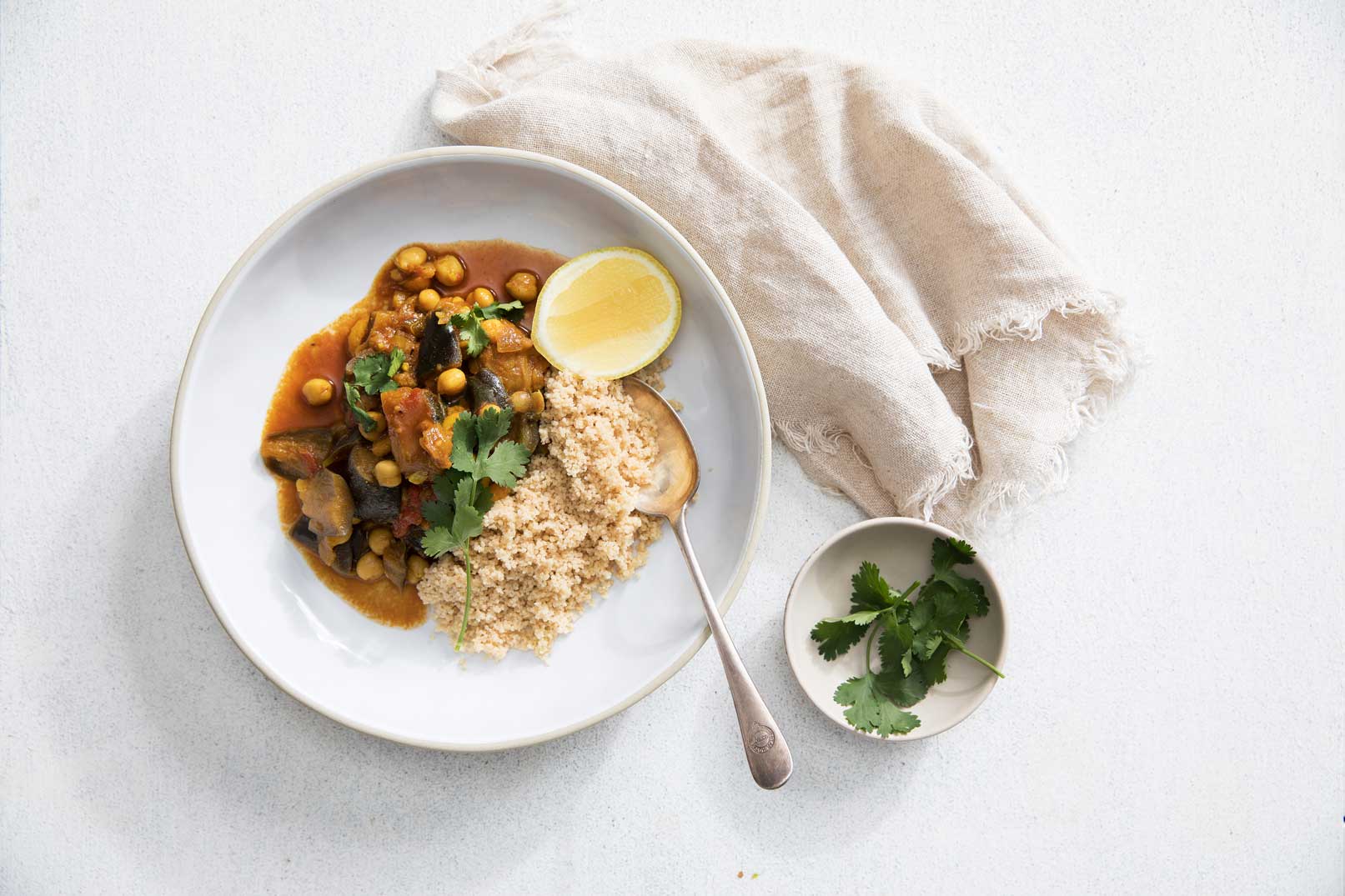 Eggplant tagine in a large white bowl with couscous, half a lemon and a silver spoon. A small bowl of coriander and a cloth napkin on the side for serving.