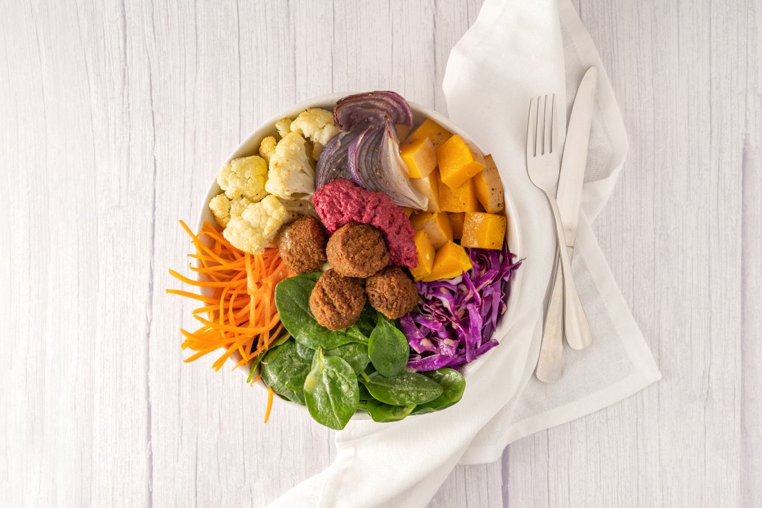 A bowl containing a colourful variety of vegetables including carrot sticks, cauliflower, green spinach, shredded red cabbage, diced pumpkin, roasted onions, beetroot hummus and falafel
