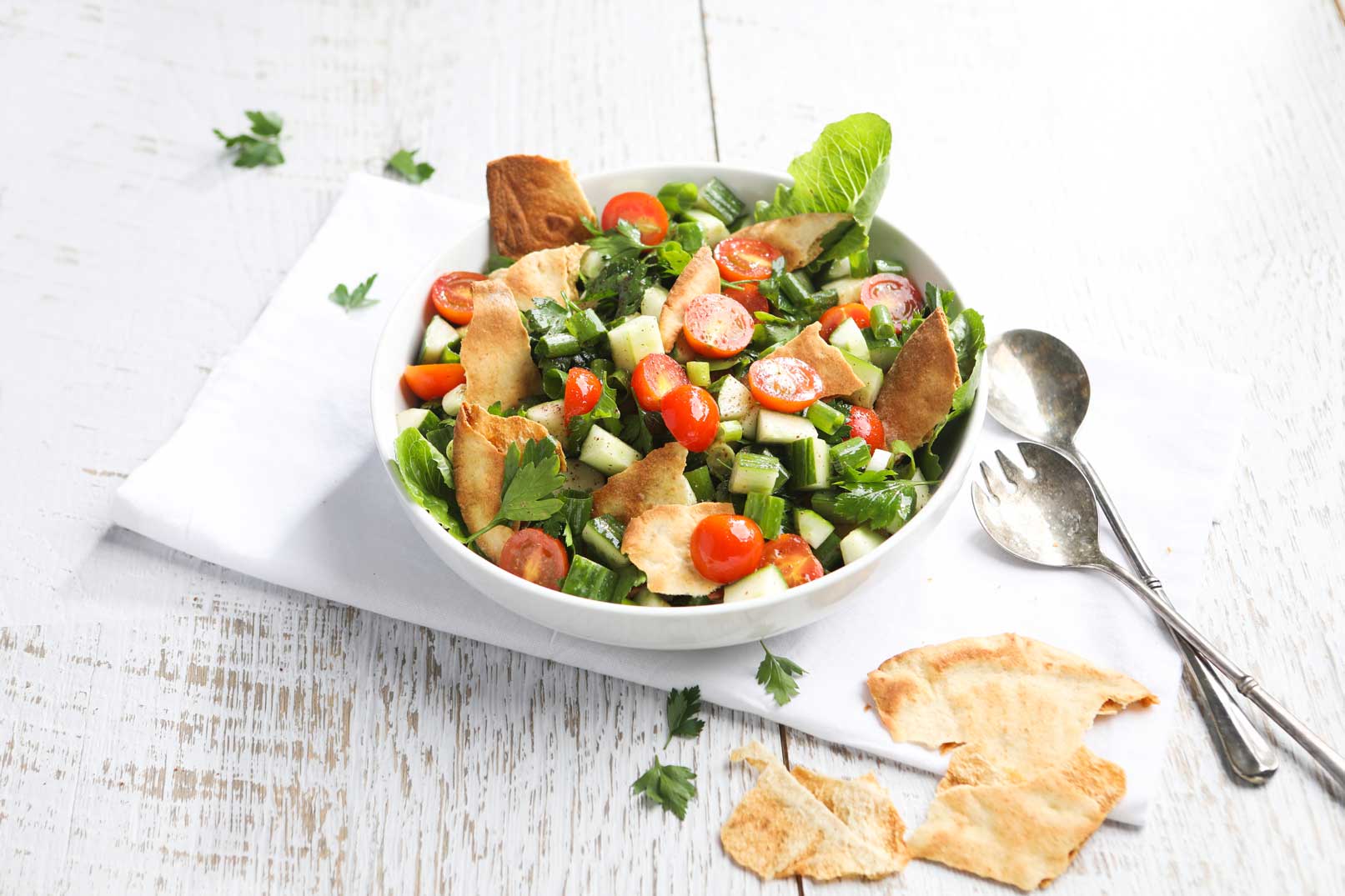 Image of fattoush salad topped with coriander in a white bowl on a white cloth napkin with serving spoons on the side.