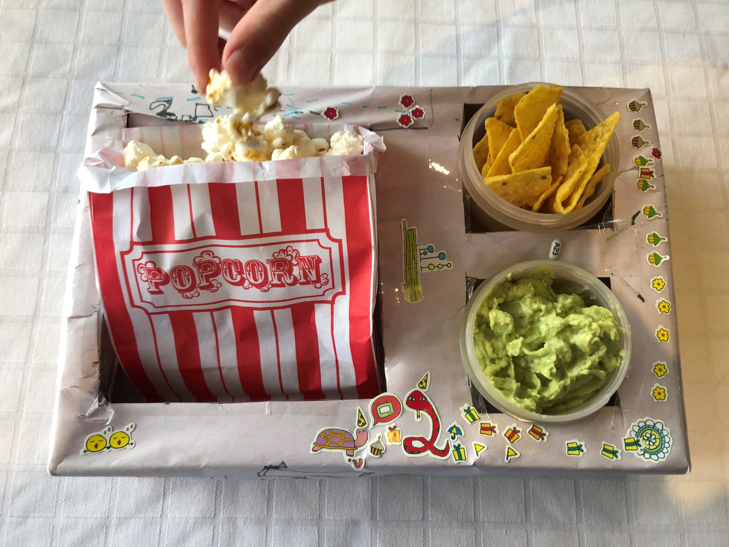 A cardboard box with cut outs for a bag of popcorn,guacamole and corn chips