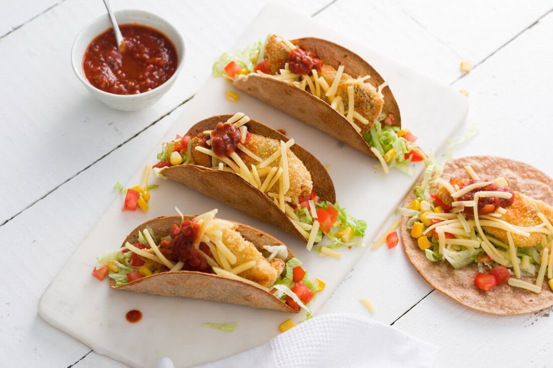 Four tacos shells containing a piece of fish with lettuce and tomato and a sprinkle of grated cheese with a small white bowl of salsa. 