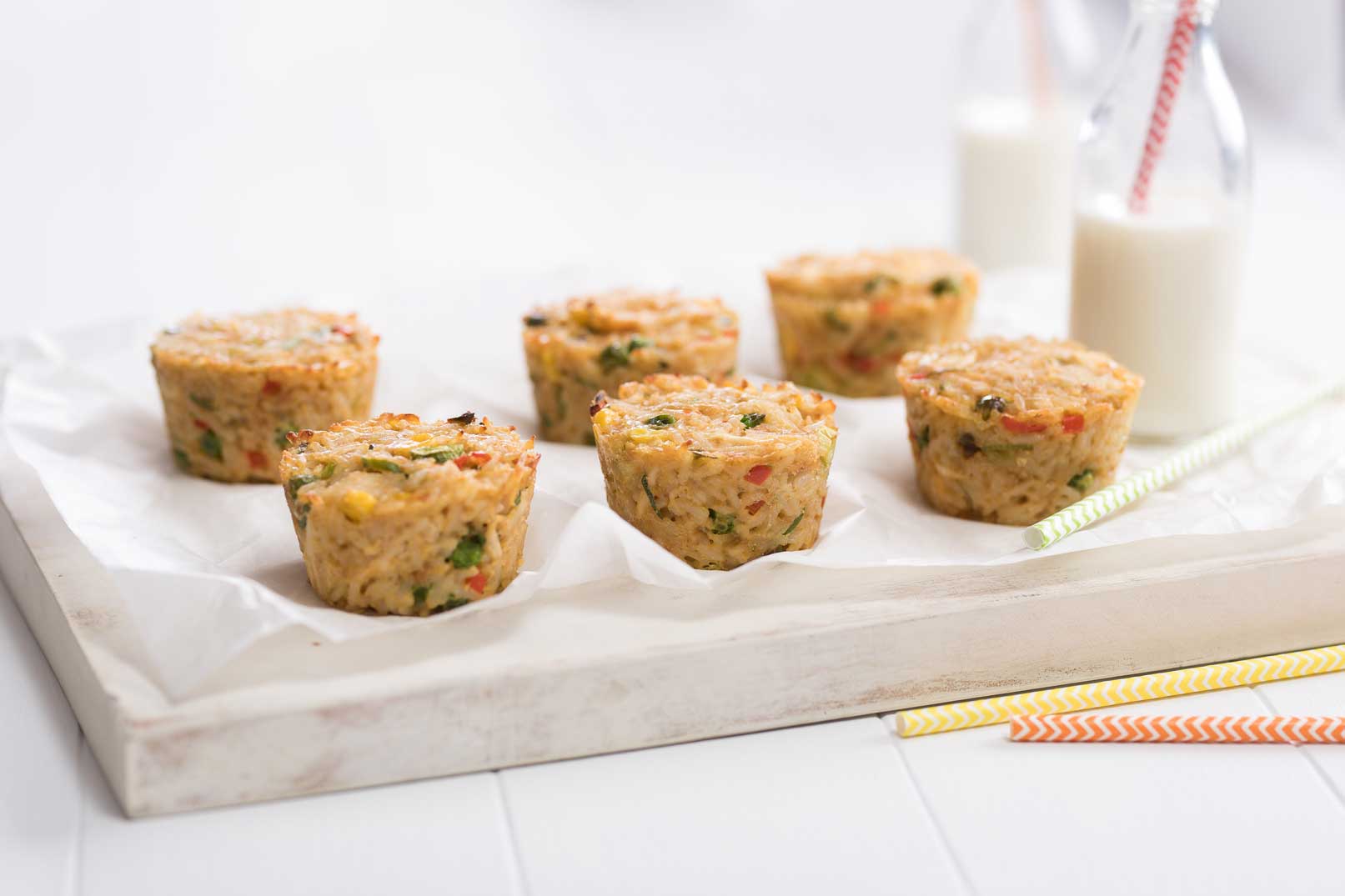 Six baked fried rice muffins on baking paper served on a white cutting board with two glass bottles of milk with straws to serve in the background.