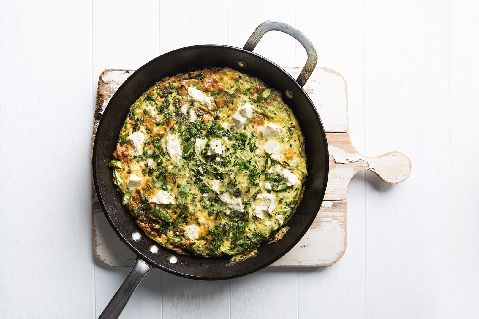 Image of a green frittata recipe cooked and served in a large round frypan sitting on a wooden cutting board
