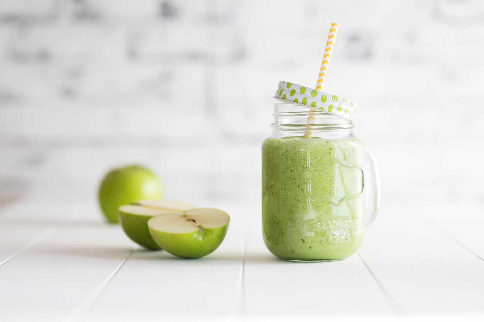 Green smoothie in a glass jug with a lid and straw and a halved green apple on the side