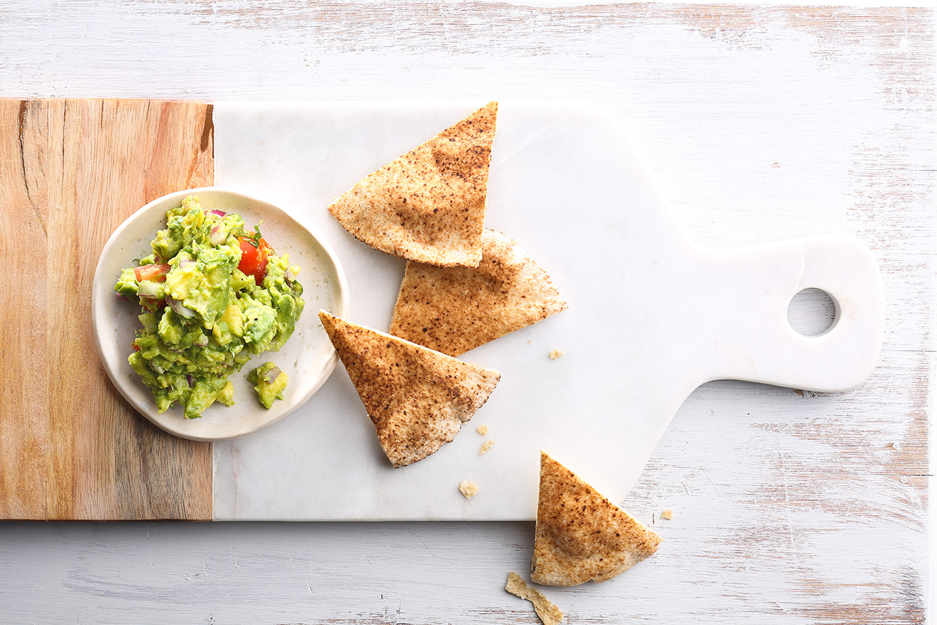 Image of a guacamole recipe in a small white bowl served on a white and wooden cutting board with baked pita triangles on the side