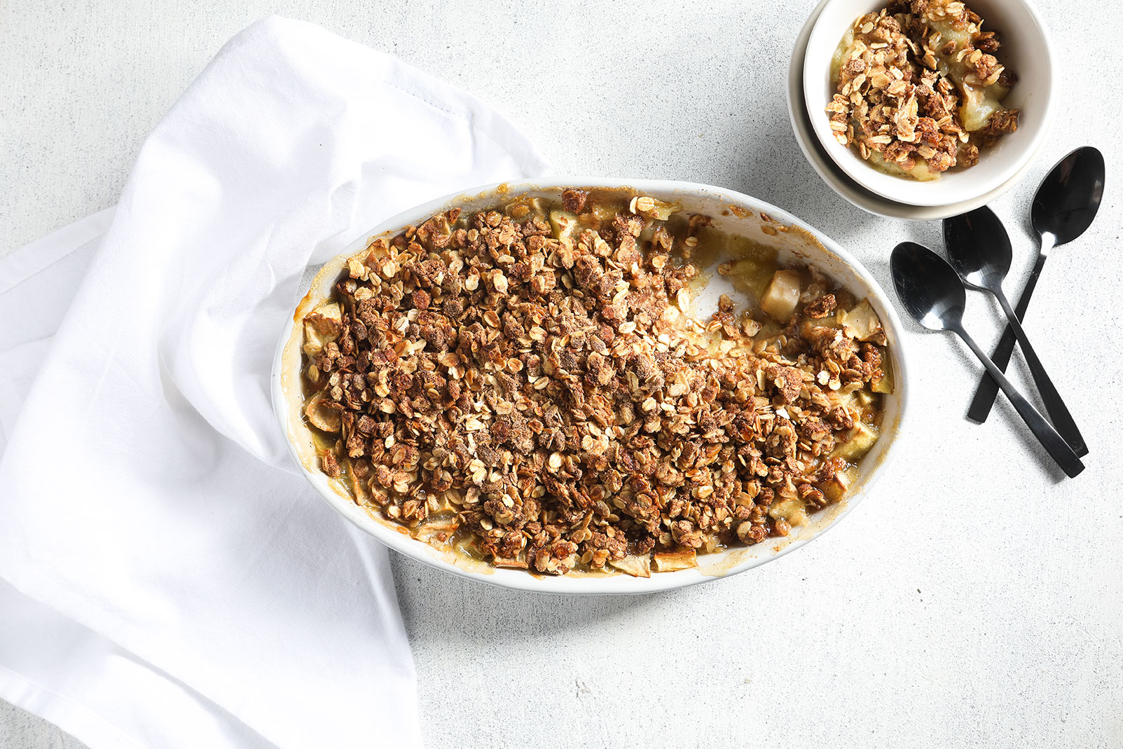 Image of baked healthy apple crumble recipe served in a large white dish with serving spoons and crumble in a small white bowl on the side