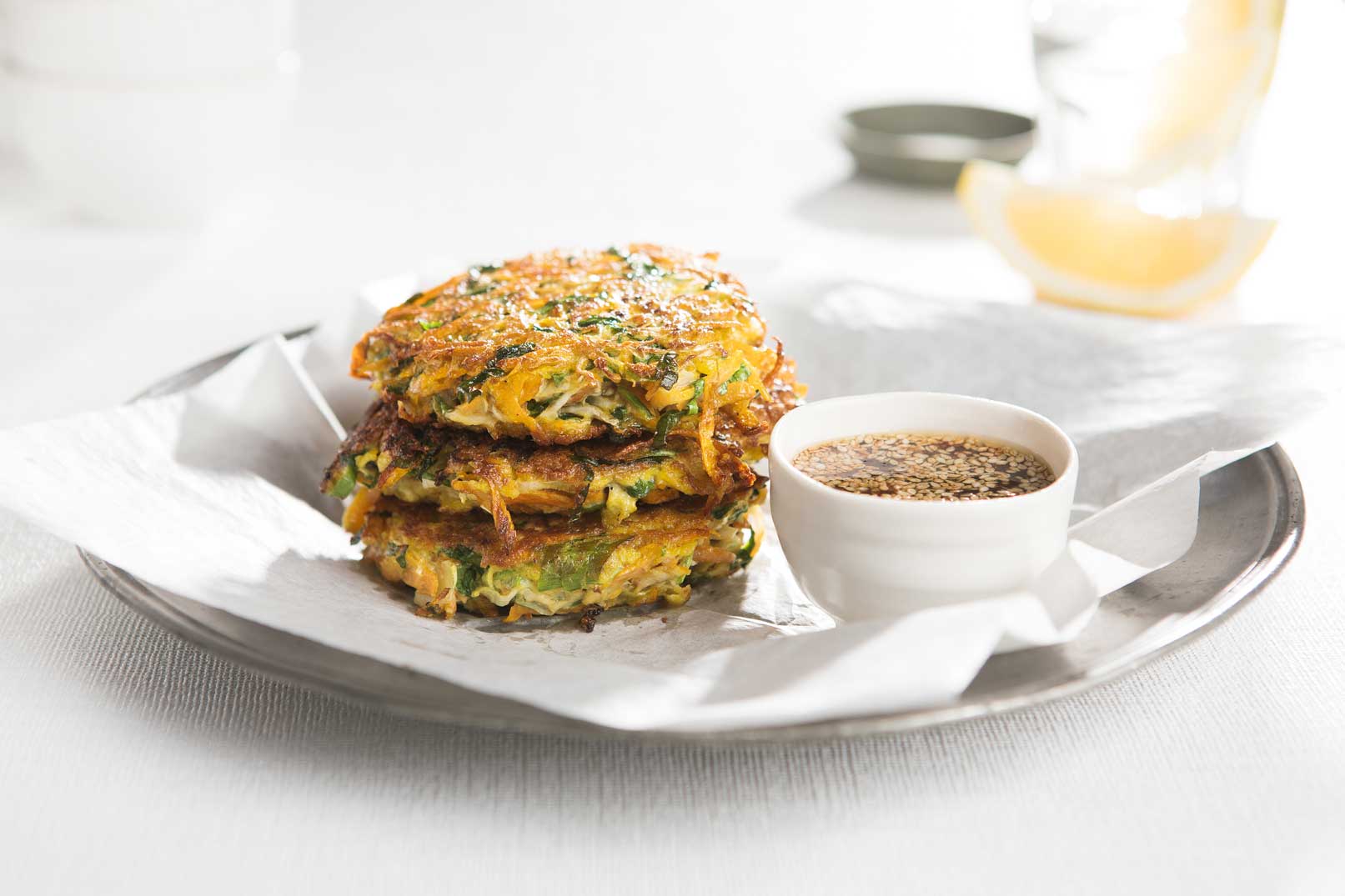 Image of three stacked vegetable pancakes on baking paper on a silver plate with a side of dipping sauce and lemon wedge.