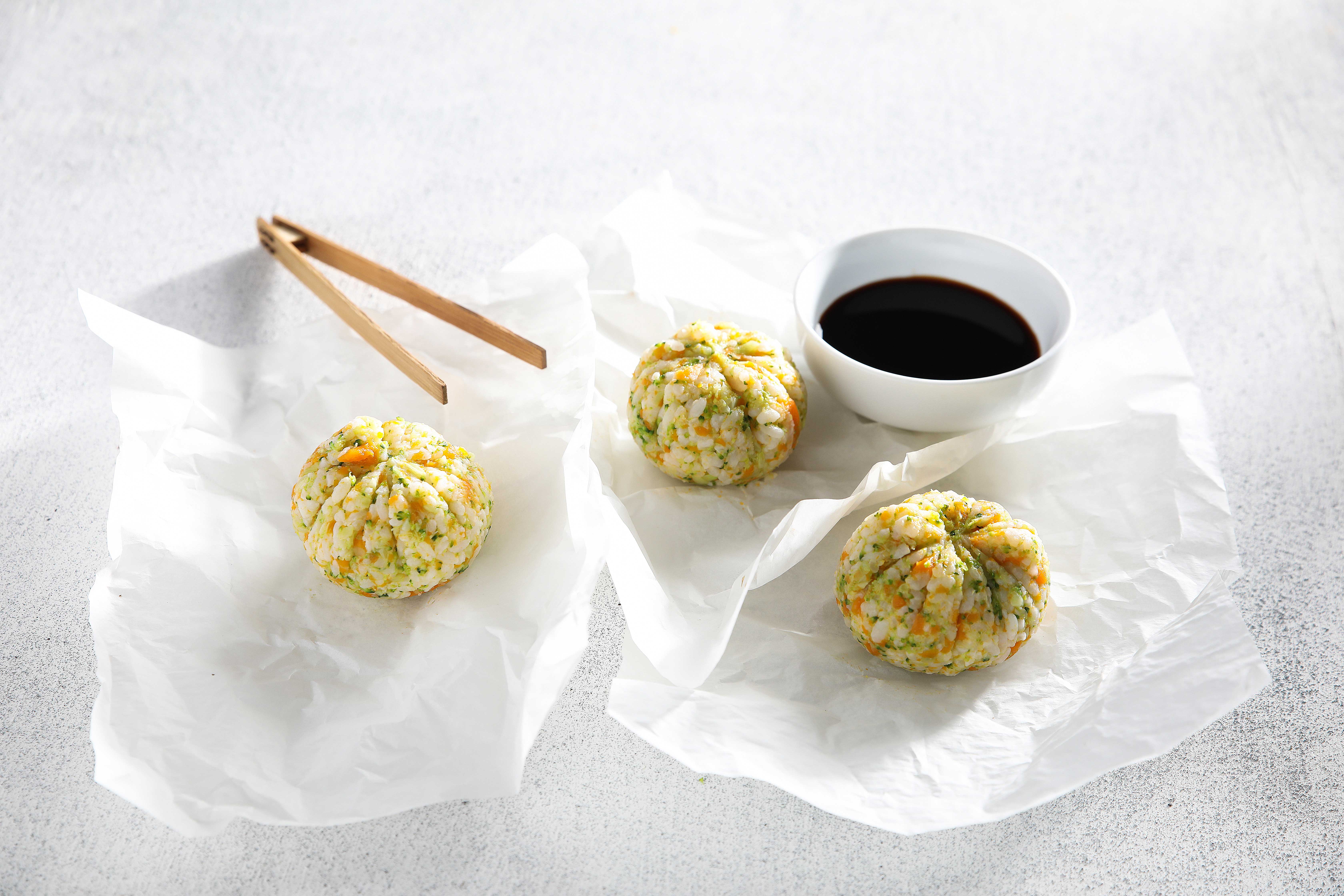 Image shot from above of three japanese rice balls okonomiyaki on baking paper with chopsticks and s small bowl of dipping sauce.