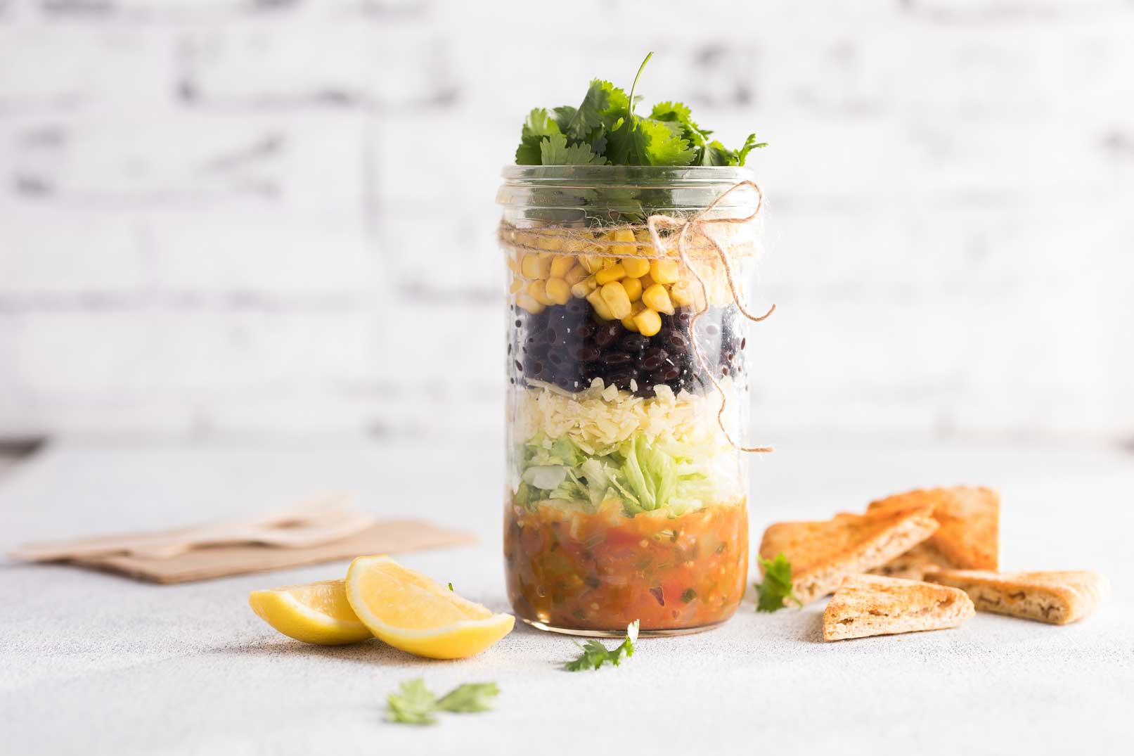 Jar nachos stacked in a tall glass jar wrapped with a string of twine. Served with pita chips, lemon wedges and a brown napkin on the side.