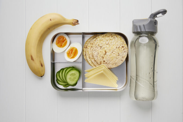 A rectangular metal lunch box containing four corn biscuits, cucumber slices, a banana, two halves of boiled egg, quarters of sliced cheese and a bottle of water