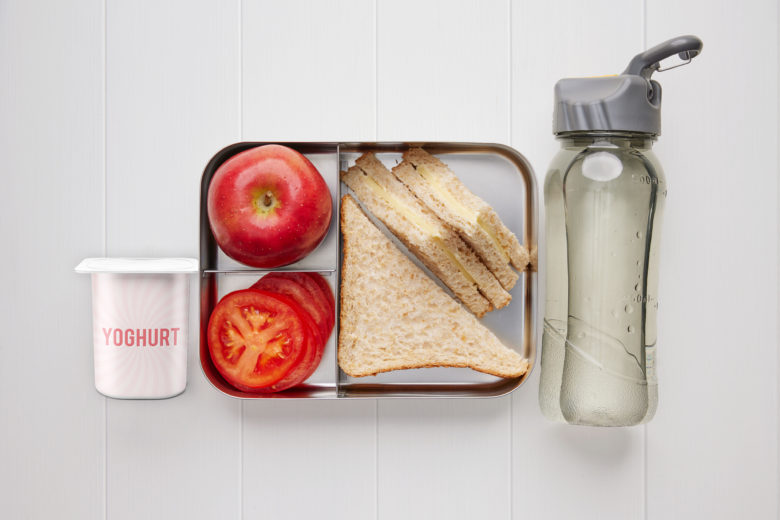 A rectangular Metal lunchbox containing cheese sandwich, tomato slices, yoghurt a red apple and a bottle of water