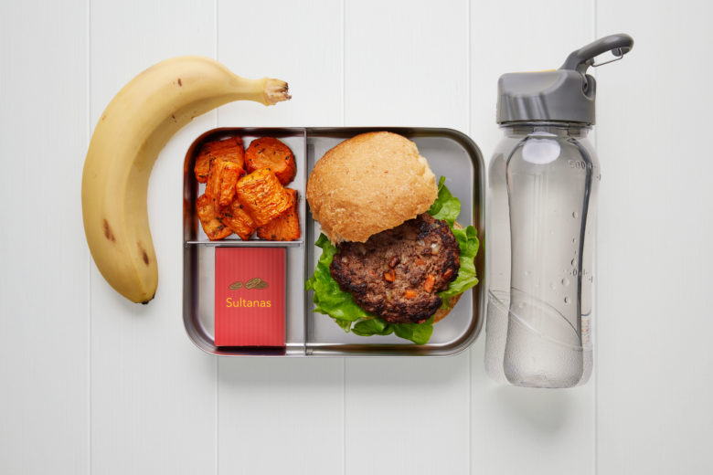 A rectangular metal lunch box containing a bread roll with a meat patty, a banana, a small carton of sultanas, some chunks of baked carrot and a bottle of water
