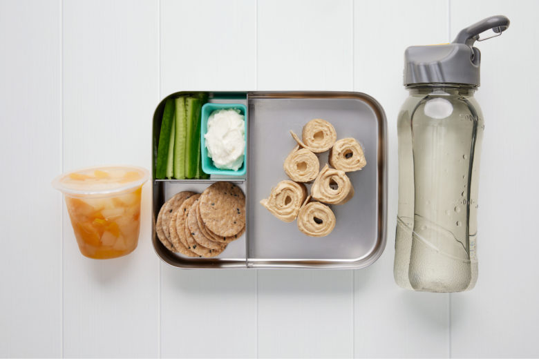 A rectangular metal lunch box containing six small pieces of a tahini wrap, rice crackers, celery and dip, a tub of tinned fruit and a bottle of water