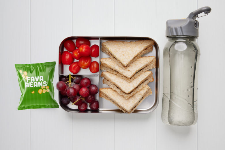 A rectangular metal lunch box containing a vegemite sandwich, red grapes, red cherry tomatoes, a packet of fava beans and a bottle of water