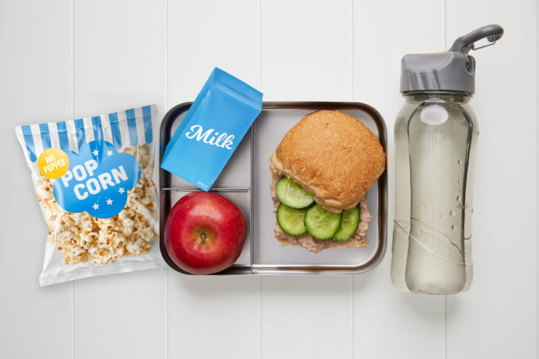 A rectangular metal lunch box containing a tuna and cucumber roll, a red apple, a carton of milk, a packet of popcorn and a bottle of water