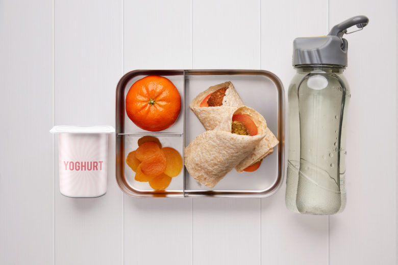 A rectangular metal lunch box containing a falafel and tomato wrap, dried apricots, a mandarin, a tub of yoghurt and a bottle of water